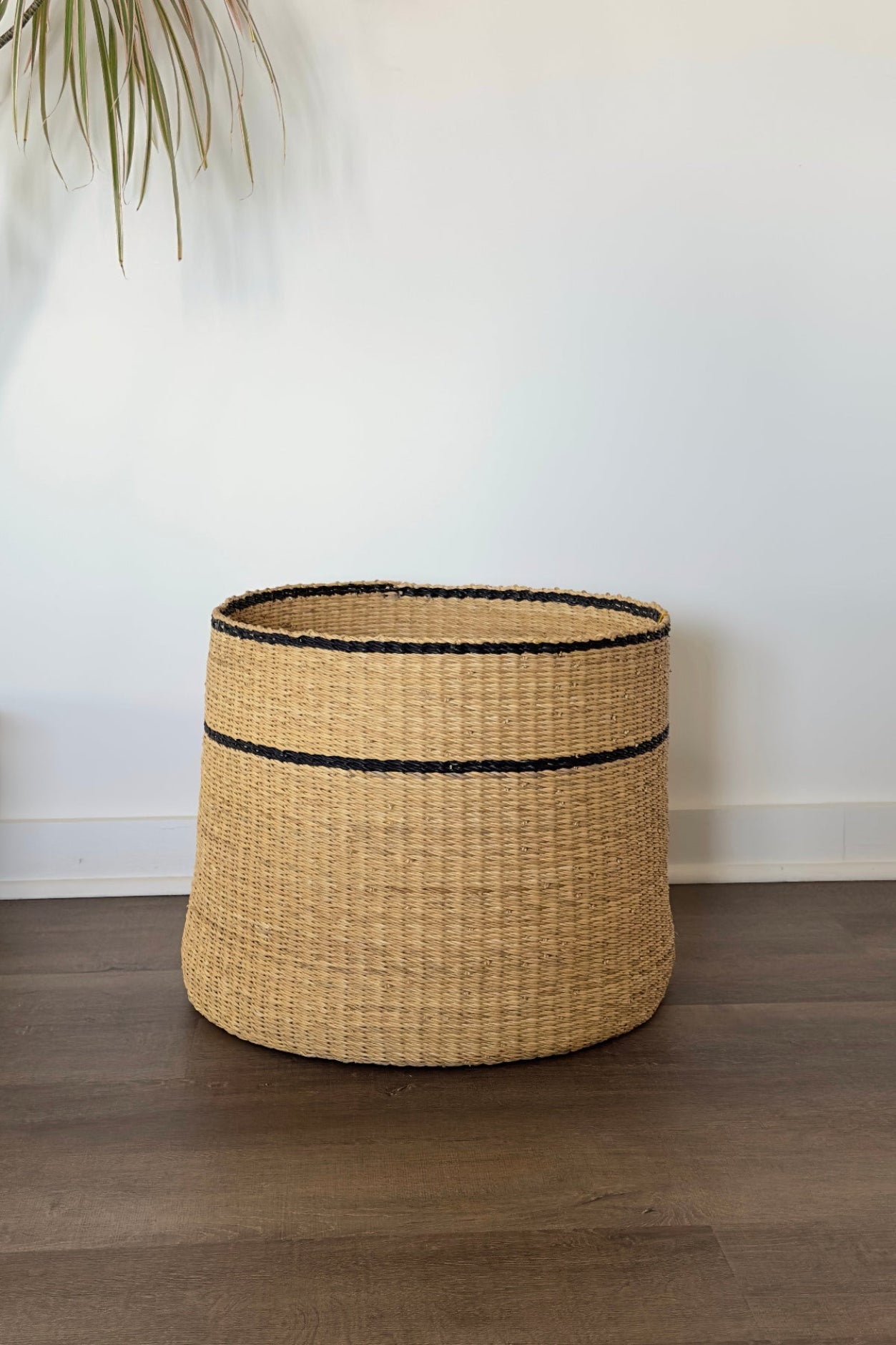 A natural woven seagrass basket with black trim, standing on a dark wood floor against a white wall.